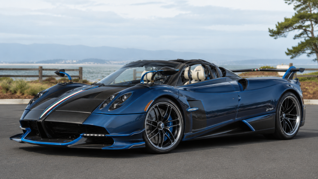 Blue Pagani Huayra Roadster parked near a waterfront with scenic mountain background.