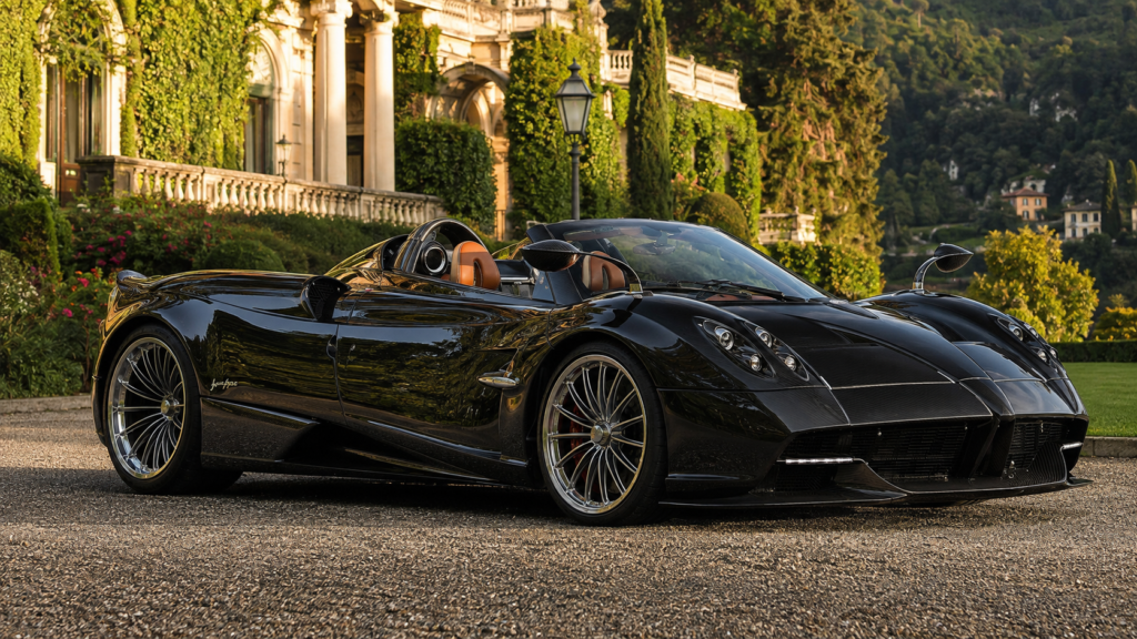 Black Pagani Huayra Roadster parked in front of a luxury villa with lush garden scenery.