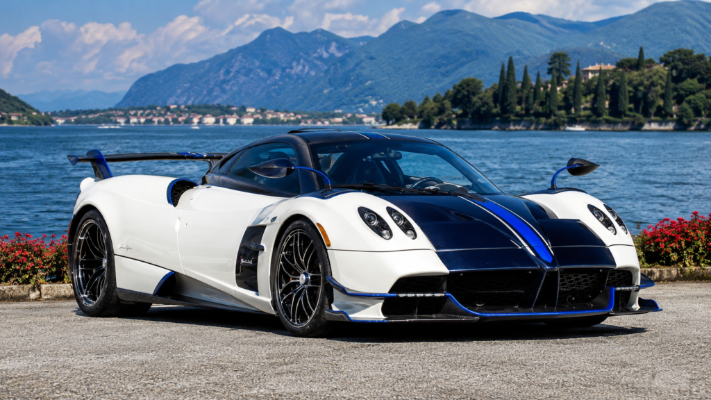 White and blue Pagani Huayra Roadster beside a picturesque lake with mountain landscape.